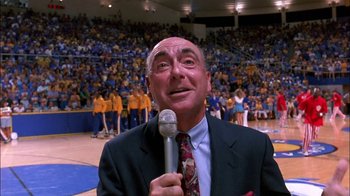 Movie still from “Blue Chips” (1994), directed by William Friedkin – A man in a suit and tie holding a microphone in front of an audience; Close Up shot, Low angle