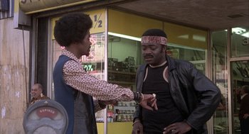 Movie still from “Blue Collar” (1978), directed by Paul Schrader – Two men standing in front of a store , one of whom is holding his hand out to the other; Medium shot, Low angle