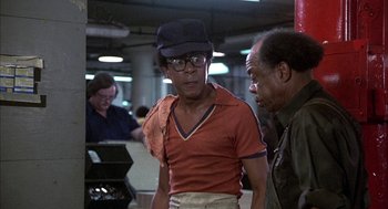 Movie still from “Blue Collar” (1978), directed by Paul Schrader – A man wearing a hat talking to another man in an airport; Medium shot, Over the shoulder angle