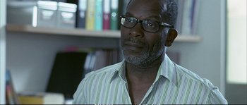 Movie still from “Boarding Gate” (2007), directed by Olivier Assayas – An older man wearing glasses and a striped shirt; Close Up shot, Over the shoulder angle