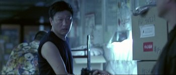 Movie still from “Boarding Gate” (2007), directed by Olivier Assayas – A man in a black tank top holding an object in a room; Medium shot, Over the shoulder angle