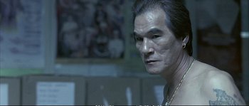 Movie still from “Boarding Gate” (2007), directed by Olivier Assayas – An older man with a long black hair and wearing a necklace; Close Up shot, Low angle