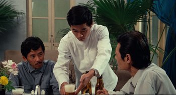 Movie still from “Boat People” (1982), directed by Ann Hui – A man pouring a beer into a glass while two other people watch; Medium shot, Over the shoulder angle