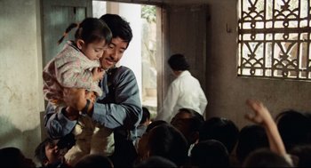 Movie still from “Boat People” (1982), directed by Ann Hui – A man holding a child in his arms in front of a group of people; Medium shot, Over the shoulder angle