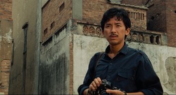Movie still from “Boat People” (1982), directed by Ann Hui – A man holding a camera while standing in front of a building; Close Up shot, Low angle