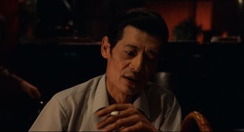 Movie still from “Boat People” (1982), directed by Ann Hui – A man sitting in front of a table with a cigarette in his mouth; Close Up shot, Over the shoulder angle