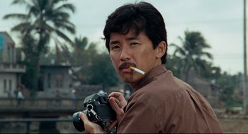 Movie still from “Boat People” (1982), directed by Ann Hui – A man holding a camera while smoking a cigarette in front of palm trees; Close Up shot, Low angle