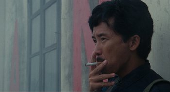 Movie still from “Boat People” (1982), directed by Ann Hui – A man is smoking a cigarette outside of a building; Close Up shot, Low angle
