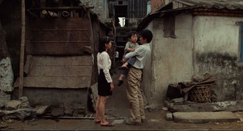 Movie still from “Boat People” (1982), directed by Ann Hui – A man holding a child while standing next to a woman; Wide shot, Over the shoulder angle