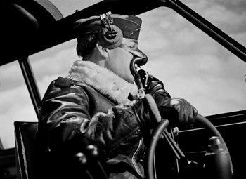 Movie still from “Bombardier” (1943), directed by Lambert Hillyer – A man in a pilot's uniform is sitting in the cockpit of an airplane; Medium shot, Low angle