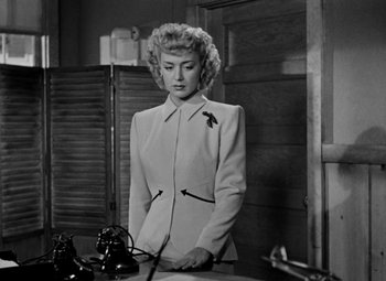 Movie still from “Bombardier” (1943), directed by Lambert Hillyer – A black and white photo of a woman standing in front of a table; Medium shot, Low angle