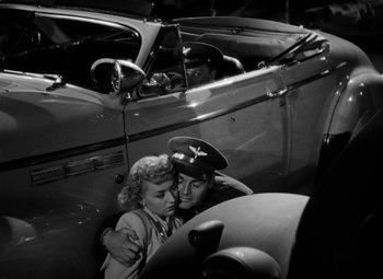 Movie still from “Bombardier” (1943), directed by Lambert Hillyer – A man and a woman hugging in front of an old car; Medium shot, Low angle