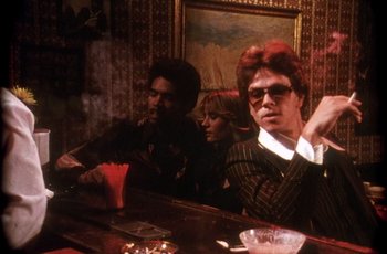Movie still from “Boogie Nights” (1997), directed by Paul Thomas Anderson – A group of people sitting at a table in front of a painting; Medium shot, Low angle