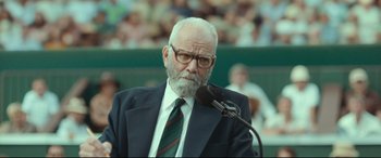 Movie still from “Borg vs. McEnroe” (2017), directed by Janus Metz – An older man in a suit and tie is speaking into a microphone in front of a crowd; Close Up shot, Over the shoulder angle