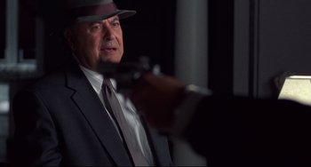 Movie still from “Bound” (1996), directed by Lilly Wachowski – An older man in a suit and a hat is holding a microphone; Close Up shot, Over the shoulder angle