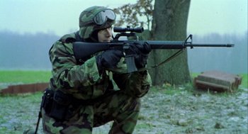 Movie still from “Bowling for Columbine” (2002), directed by Michael Moore – A man in camouflage with a rifle in front of a tree; Medium shot, High angle