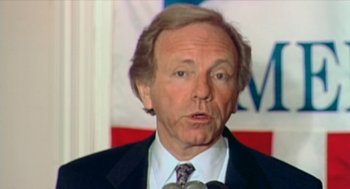 Movie still from “Bowling for Columbine” (2002), directed by Michael Moore – A man in a suit and tie speaking into a microphone in front of an american flag; Close Up shot, Low angle