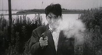 Movie still from “Bowling for Columbine” (2002), directed by Michael Moore – A man holding a gun while standing next to a body of water; Medium shot, Low angle