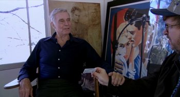 Movie still from “Bowling for Columbine” (2002), directed by Michael Moore – An older man sitting in front of an older woman holding a business card; Medium shot, Over the shoulder angle
