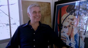 Movie still from “Bowling for Columbine” (2002), directed by Michael Moore – An older man is smiling in front of a painting; Medium shot, Over the shoulder angle