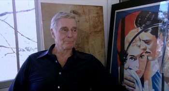 Movie still from “Bowling for Columbine” (2002), directed by Michael Moore – An older man standing in front of a painting; Close Up shot, Over the shoulder angle