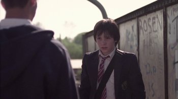 Movie still from “Boy A” (2007), directed by John Crowley – A boy in a suit and tie standing in front of a wall; Medium shot, Over the shoulder angle