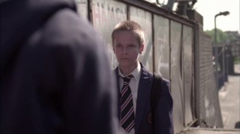 Movie still from “Boy A” (2007), directed by John Crowley – A boy in a suit and tie standing in front of a wall; Medium shot, Over the shoulder angle
