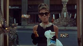 Movie still from “Breakfast at Tiffany's” (1961), directed by Blake Edwards – A woman in a black dress eating a donut and drinking coffee; Medium shot, Over the shoulder angle