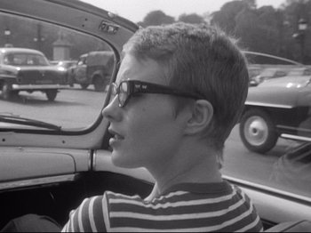 Movie still from “Breathless” (1960), directed by Jean-Luc Godard – A young boy wearing glasses sitting in the driver's seat of a car; Close Up shot, Over the shoulder angle