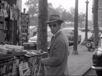 Movie still from “Breathless” (1960), directed by Jean-Luc Godard – A black and white photo of a man standing in front of a newspaper stand; Medium shot, High angle