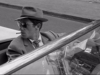 Movie still from “Breathless” (1960), directed by Jean-Luc Godard – A black and white photo of a man in a suit and hat; Close Up shot, Over the shoulder angle