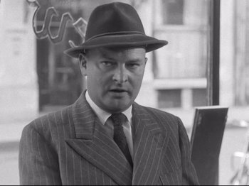 Movie still from “Breathless” (1960), directed by Jean-Luc Godard – A man in a suit and hat is standing in front of a building; Close Up shot, Low angle