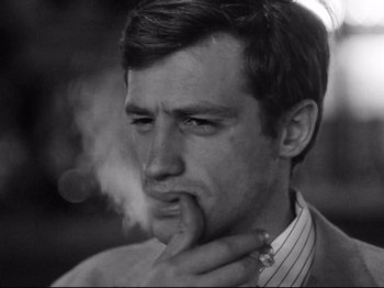Movie still from “Breathless” (1960), directed by Jean-Luc Godard – A black and white photo of a man smoking a cigarette; Close Up shot, High angle