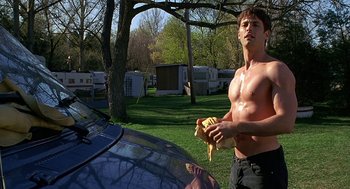 Movie still from “Bride of Chucky” (1998), directed by Ronny Yu – A shirtless man holding a banana next to a car; Medium shot, Over the shoulder angle