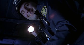 Movie still from “Bride of Chucky” (1998), directed by Ronny Yu – A police officer holding a flashlight and smiling for the camera; Close Up shot, Low angle