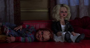 Movie still from “Bride of Chucky” (1998), directed by Ronny Yu – A doll laying next to another doll on a red couch; Medium shot, Overhead angle