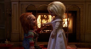 Movie still from “Bride of Chucky” (1998), directed by Ronny Yu – A couple of dolls that are in front of a fireplace; Medium shot, Over the shoulder angle