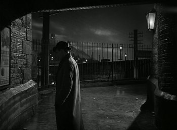 Movie still from “Brief Encounter” (1945), directed by David Lean – A black and white photo of a man in a trench coat; Wide shot, Over the shoulder angle
