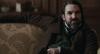 Movie still from “Bright Star” (2009), directed by Jane Campion – A man sitting on top of a couch holding a book; Medium shot, Over the shoulder angle