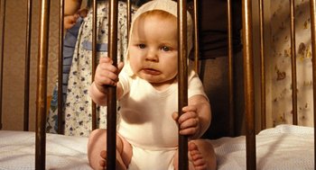 Movie still from “Bronson” (2008), directed by Nicolas Winding Refn – A baby in a white onesie is sitting in a crib; Medium shot, High angle