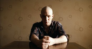 Movie still from “Bronson” (2008), directed by Nicolas Winding Refn – A bald man sitting at a table with his arms crossed; Medium shot, High angle