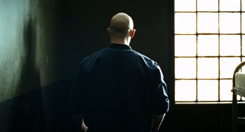 Movie still from “Bronson” (2008), directed by Nicolas Winding Refn – A man standing in front of a window in a dark room; Medium shot, Low angle