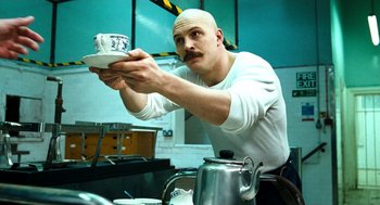 Movie still from “Bronson” (2008), directed by Nicolas Winding Refn – A bald man with a mustache holding a coffee cup; Medium shot, Low angle