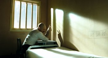 Movie still from “Bronson” (2008), directed by Nicolas Winding Refn – A man sitting on top of a bed next to a window; Medium shot, High angle