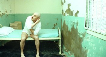 Movie still from “Bronson” (2008), directed by Nicolas Winding Refn – A bald man sitting on a bed in a room; Medium shot, High angle