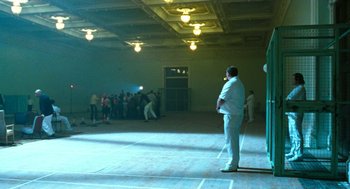 Movie still from “Bronson” (2008), directed by Nicolas Winding Refn – A man standing in the middle of a room with a crowd of onlookers; Wide shot, Low angle
