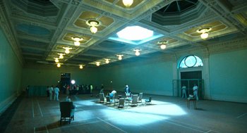 Movie still from “Bronson” (2008), directed by Nicolas Winding Refn – People are gathered in a large room with a light fixture; Extreme Wide shot, High angle