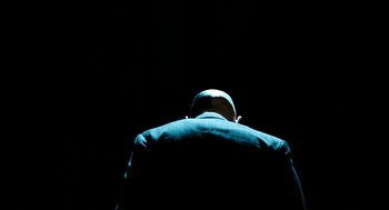 Movie still from “Bronson” (2008), directed by Nicolas Winding Refn – A man in a suit and hat is sitting in the dark; Medium shot, Low angle