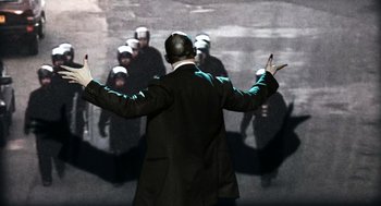Movie still from “Bronson” (2008), directed by Nicolas Winding Refn – A man in a black suit and a helmet is standing in front of a group of people; Medium shot, Over the shoulder angle