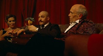 Movie still from “Bronson” (2008), directed by Nicolas Winding Refn – Two men sitting on a red couch with one holding an object; Medium shot, Low angle